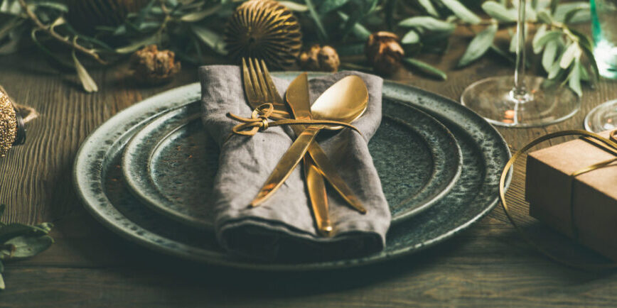 Christmas or New Years celebration party table setting. Plates, golden cutlery, glasses, gift box, festive branch decoration, candles, gliterring toys over rustic wooden table background