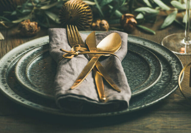 Christmas or New Years celebration party table setting. Plates, golden cutlery, glasses, gift box, festive branch decoration, candles, gliterring toys over rustic wooden table background