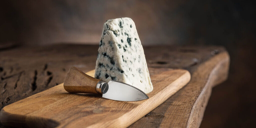 Piece of blue cheese and cheese knife on the wooden board. Dark background.